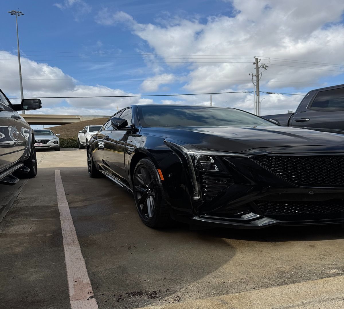 A glossy black Cadillac CT5-V Blackwing parked in a paved lot under a blue sky with scattered white clouds.