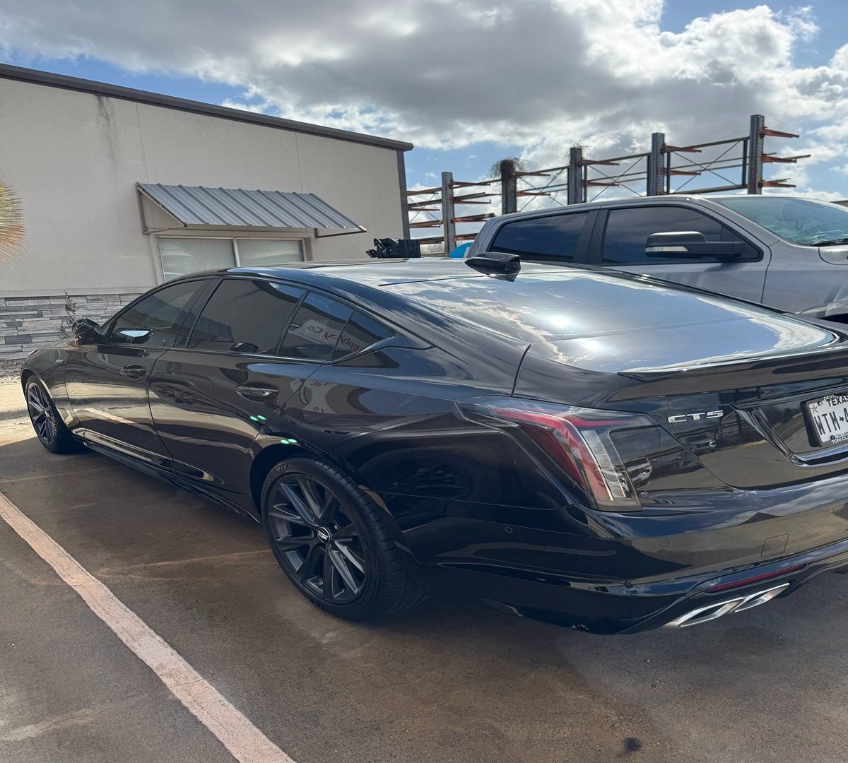 A black Cadillac CT5 sedan parked in a paved lot next to a light-colored building and a silver pickup truck.