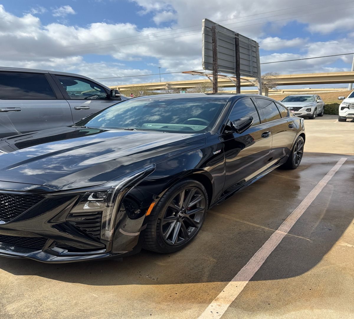 A sleek black Cadillac CT5 sedan parked in a sunlit, paved outdoor lot under a cloudy blue sky.