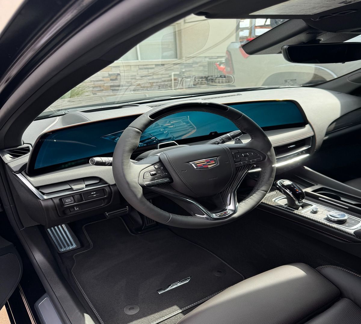 Interior view of a Cadillac vehicle showing the dashboard, curved digital display, steering wheel, and black floor mats.