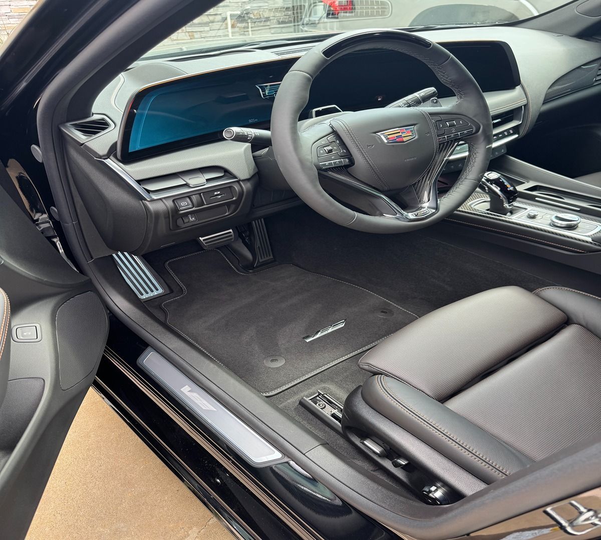 The interior of a modern Cadillac vehicle, showing a black leather seat, steering wheel, and digital dashboard.
