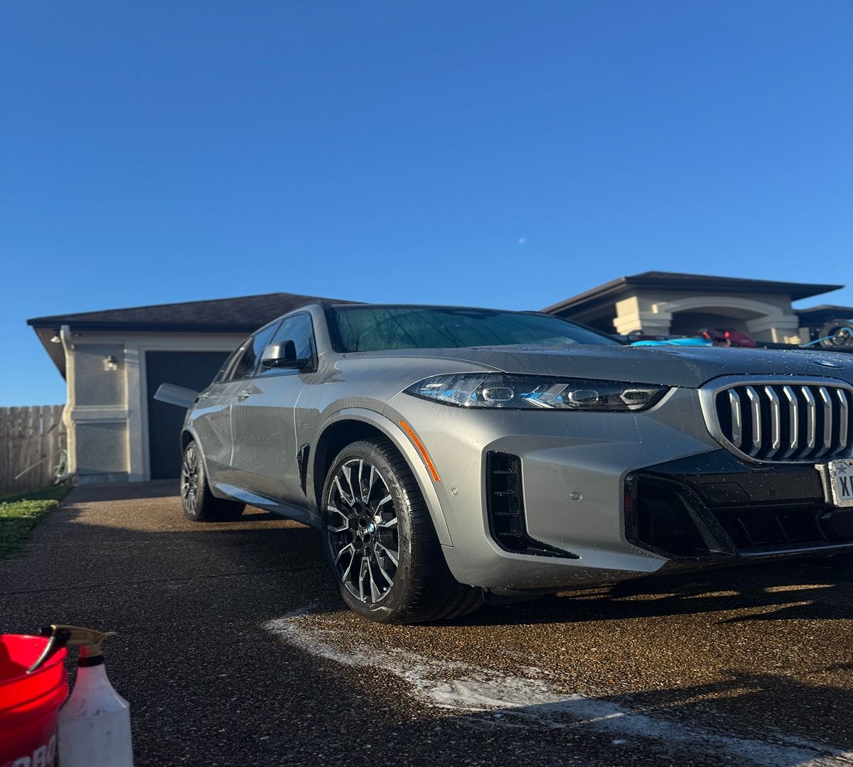 A silver BMW SUV parked in a residential driveway on a clear, sunny day, with cleaning supplies visible nearby.