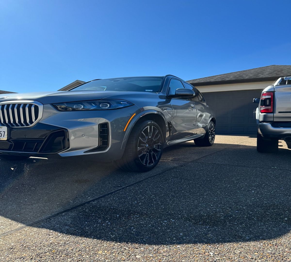A grey BMW SUV parked in a driveway next to a truck on a sunny day.