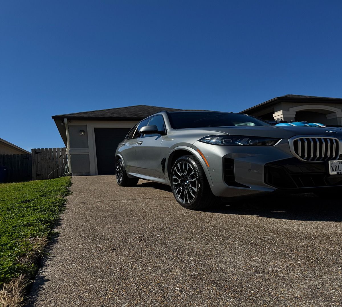 A gray BMW parked on a concrete driveway in front of a house under a clear blue sky.