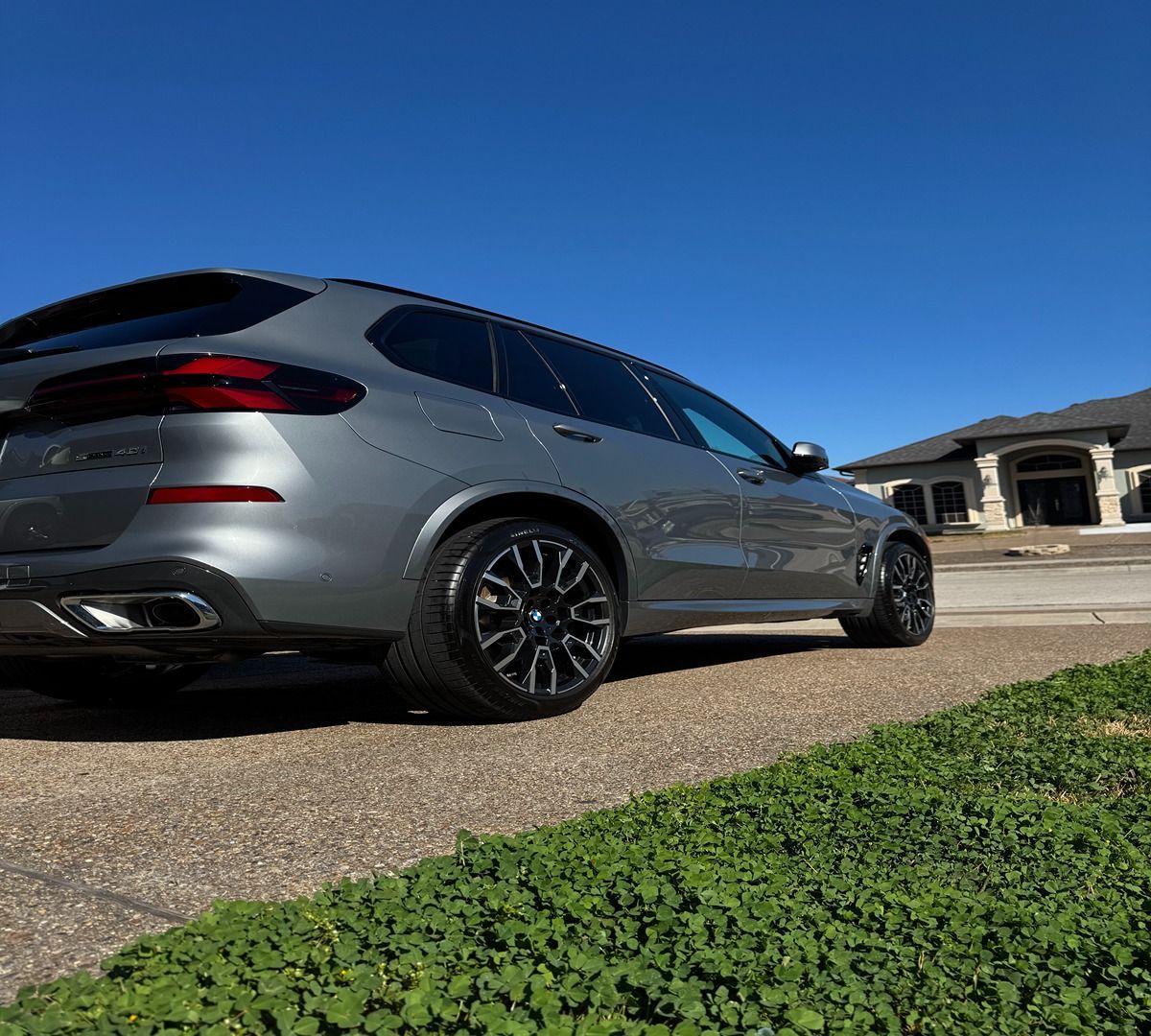 A gray BMW SUV parked on a paved driveway in front of a house under a clear blue sky.