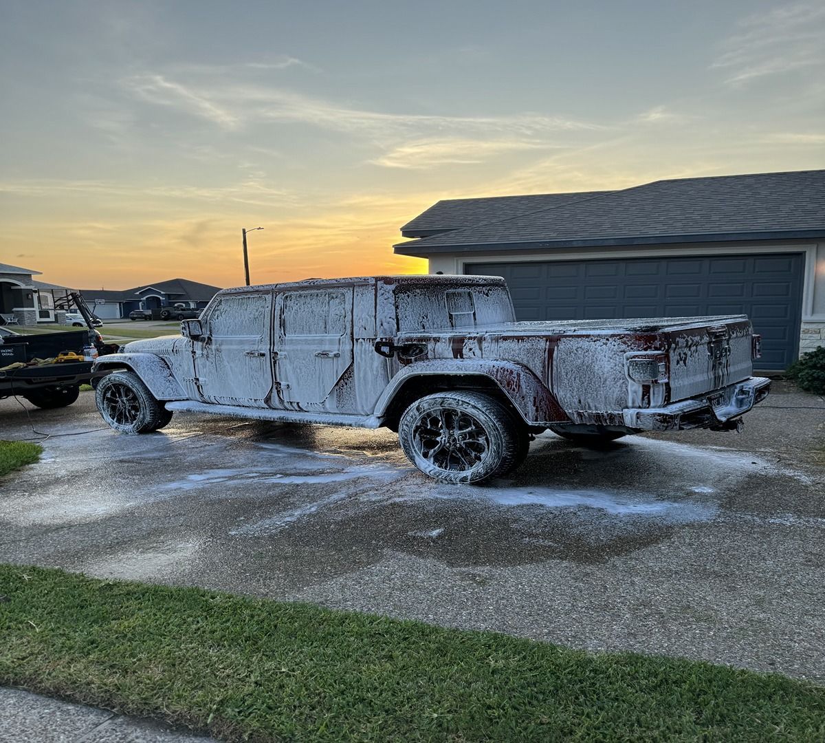 A Jeep Gladiator pickup truck covered in thick white soap suds, parked on a driveway at sunset.