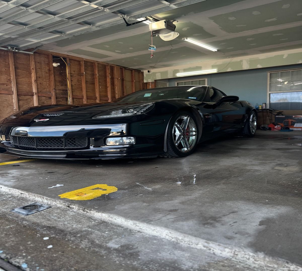 A sleek black Corvette parked inside a residential garage with unfinished wooden walls and concrete flooring.
