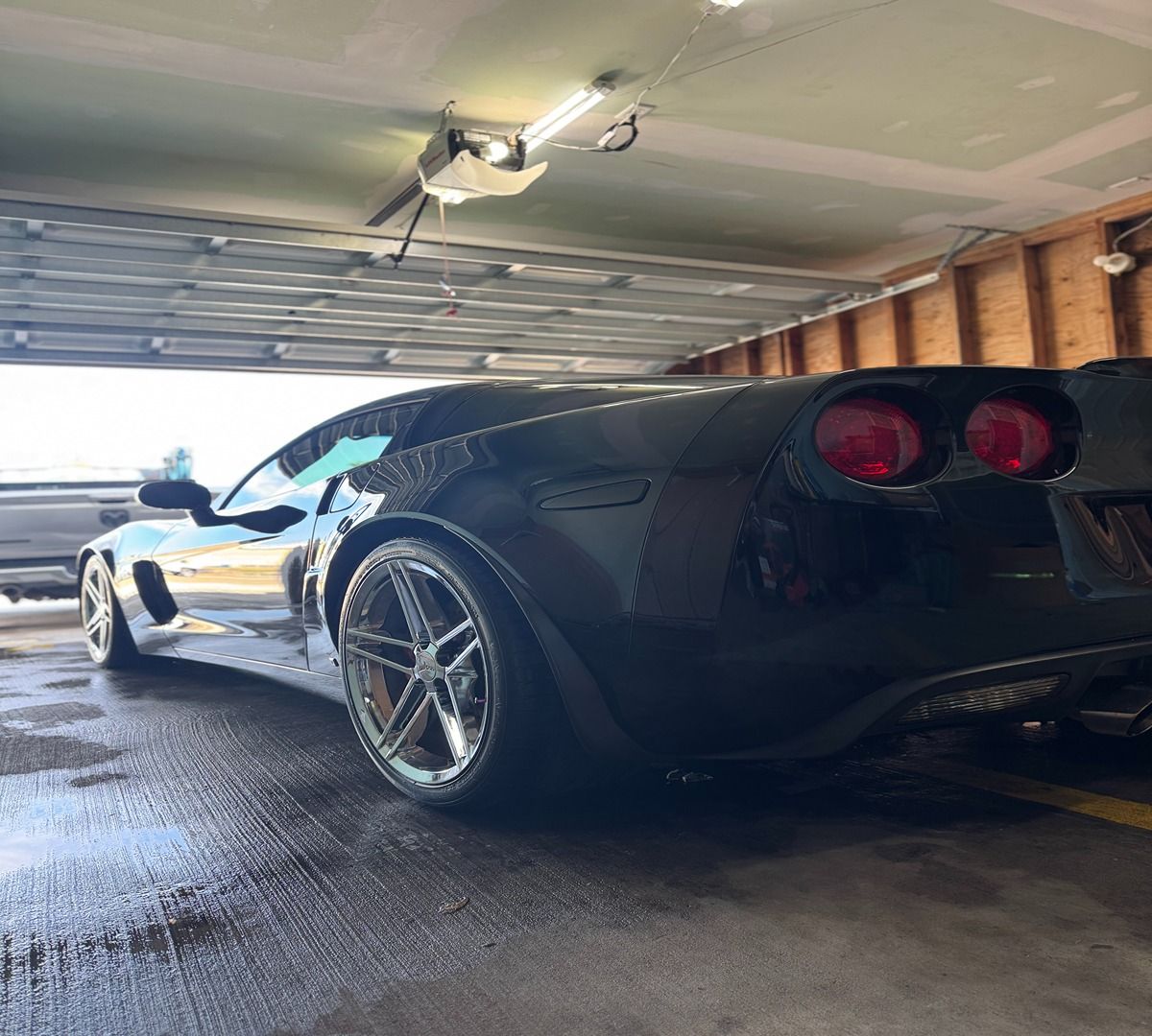 A sleek, black Chevrolet Corvette parked inside a residential garage, viewed from the rear three-quarter angle.