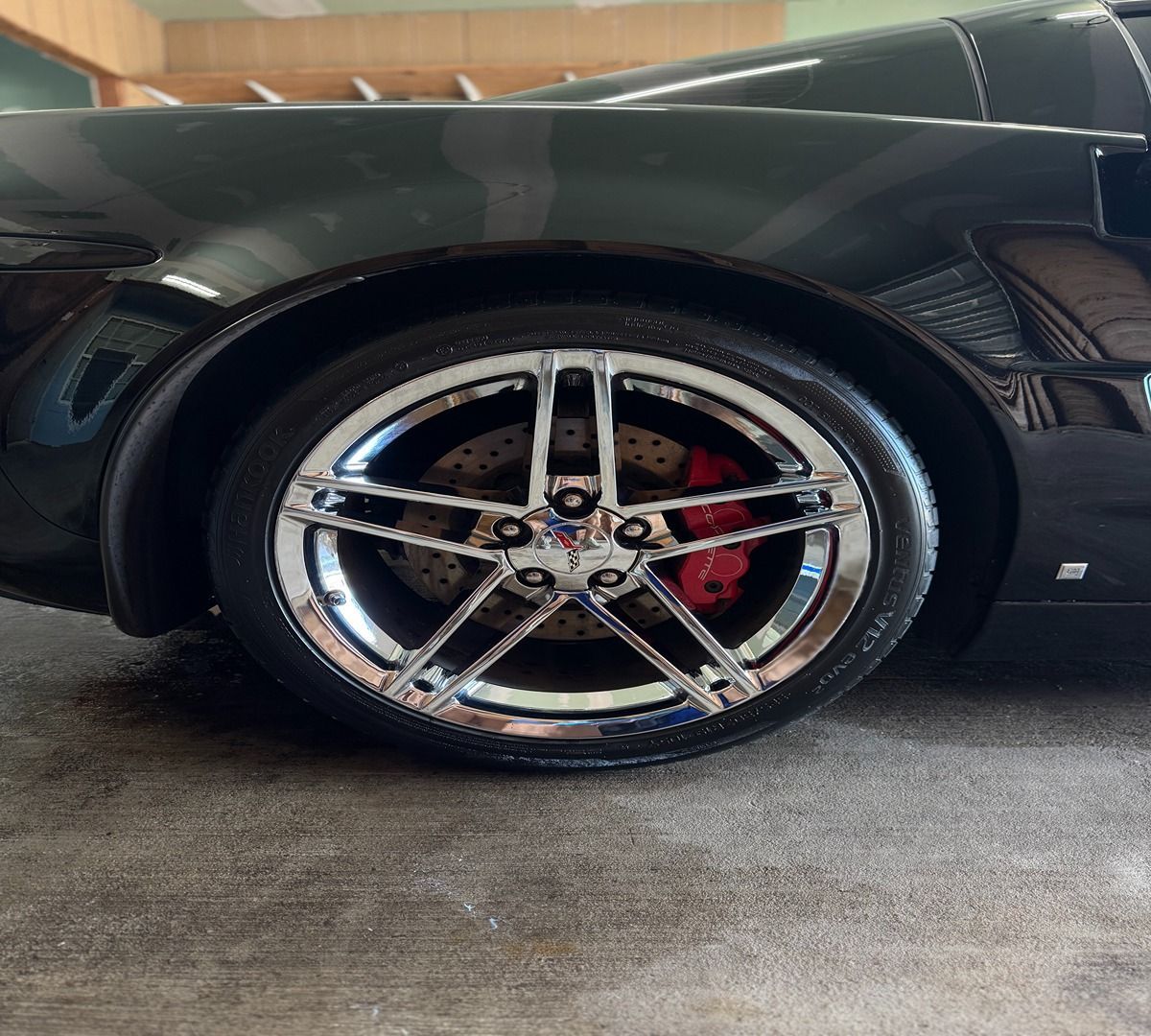 A close-up, side profile of a black Corvette showing a chrome five-spoke wheel with red brake calipers on a concrete floor.