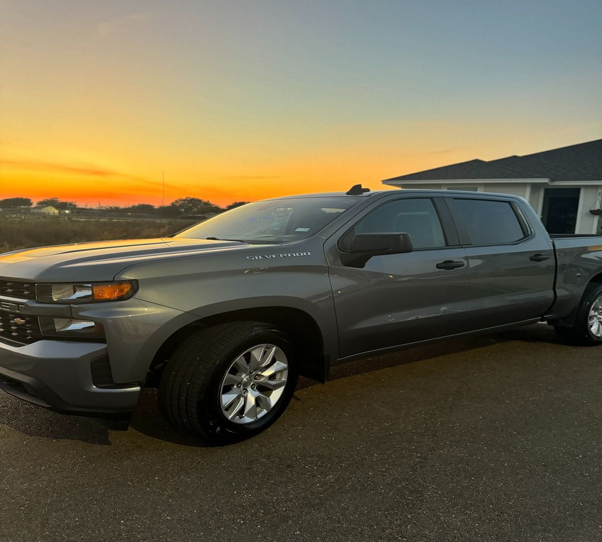 A grey Chevrolet Silverado pickup truck parked outside a home during a vibrant orange and yellow sunset.