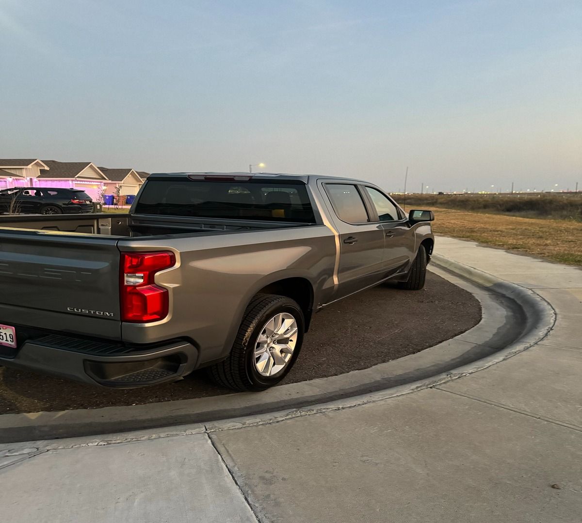 A dark gray Chevrolet pickup truck parked on a concrete curve near a suburban housing development at sunset.