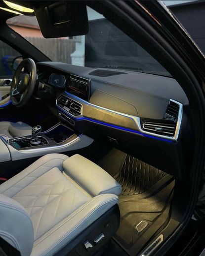 The driver-side interior of a modern BMW featuring light-grey patterned leather seats, black dashboard, and blue ambient light.