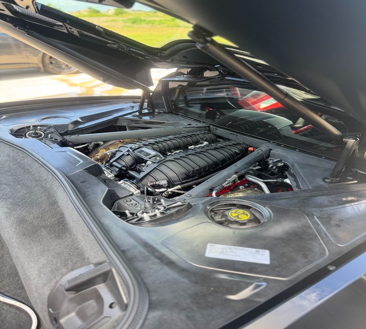 An open engine compartment of a mid-engine sports car, showcasing the black intake manifold and mechanical components.