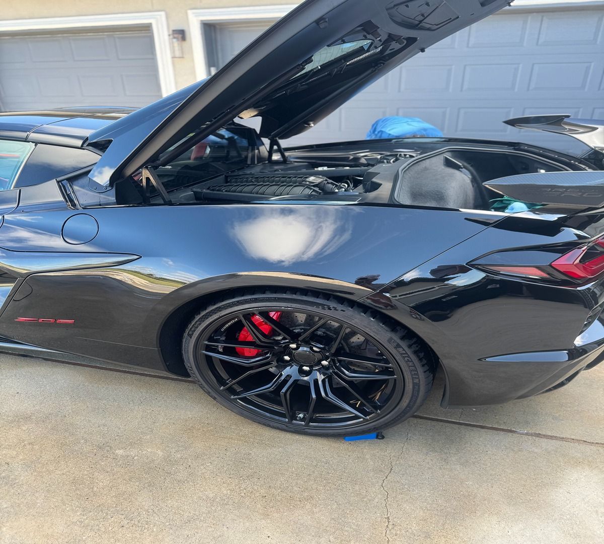 A black Chevrolet Corvette C8 with its engine cover open, parked on a concrete driveway in front of a garage.