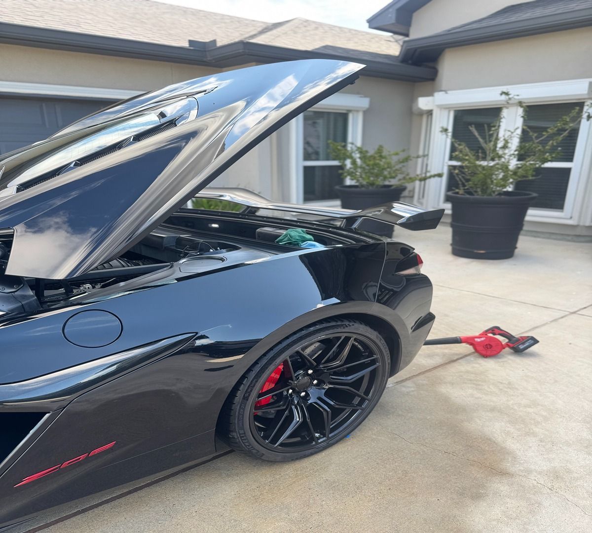 A black sports car with its rear engine cover open, parked on a concrete driveway near a house.