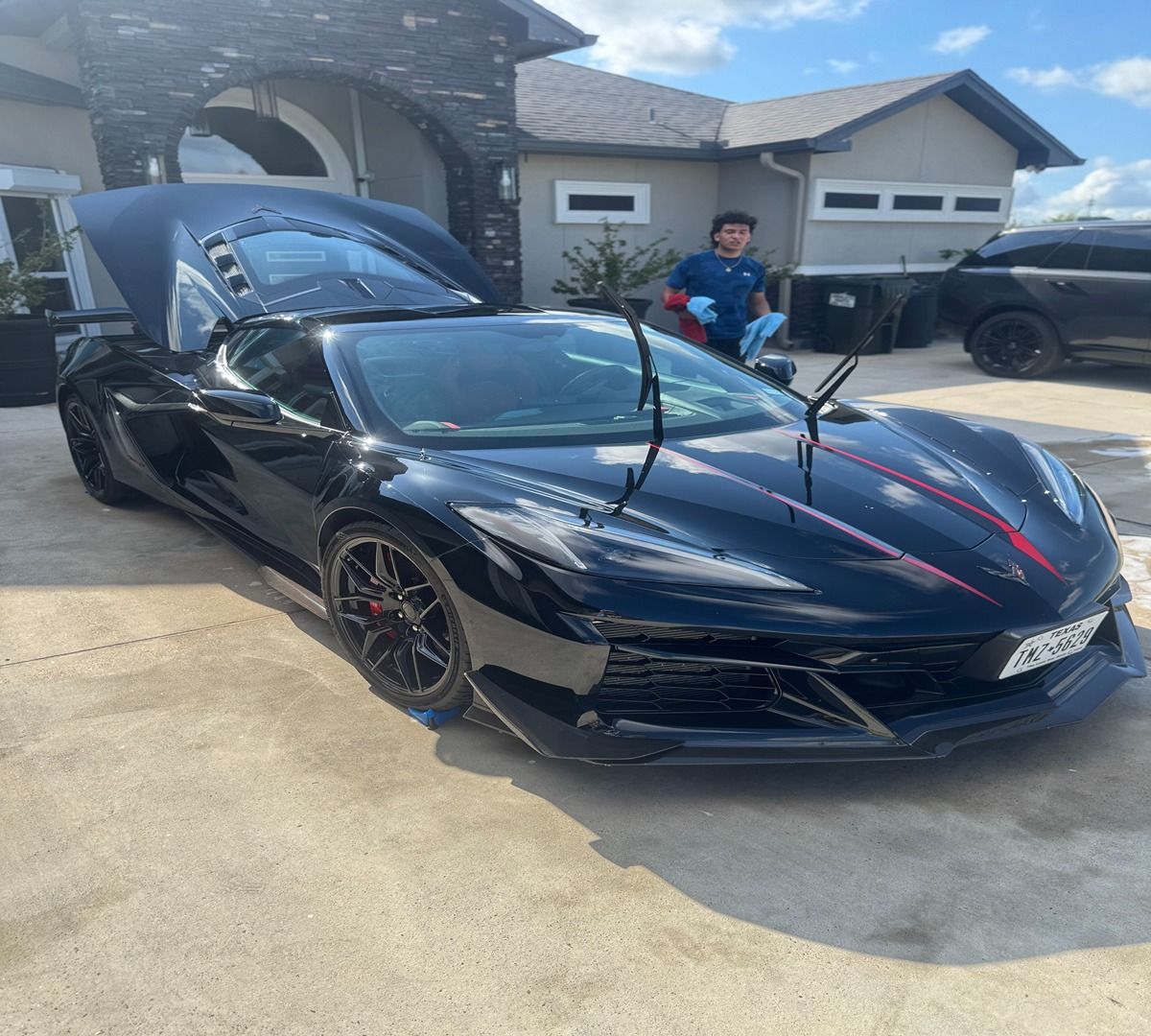 A person stands behind a sleek, black sports car with its engine hood open, parked in front of a house.