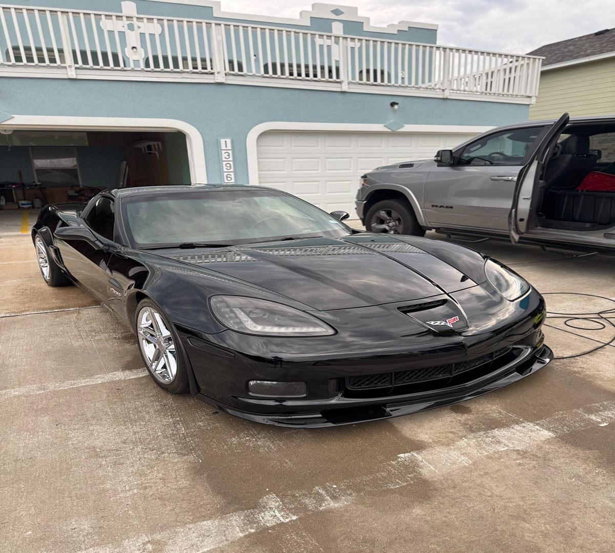 A black Chevrolet Corvette parked on a concrete driveway in front of a house with a grey pickup truck nearby.