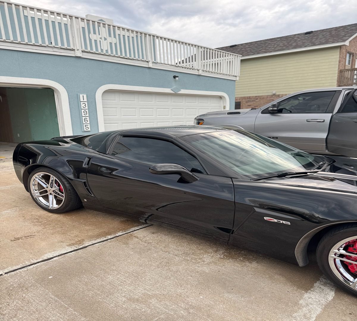 A black Corvette parked on a concrete driveway in front of a house with a white garage door and a gray truck.