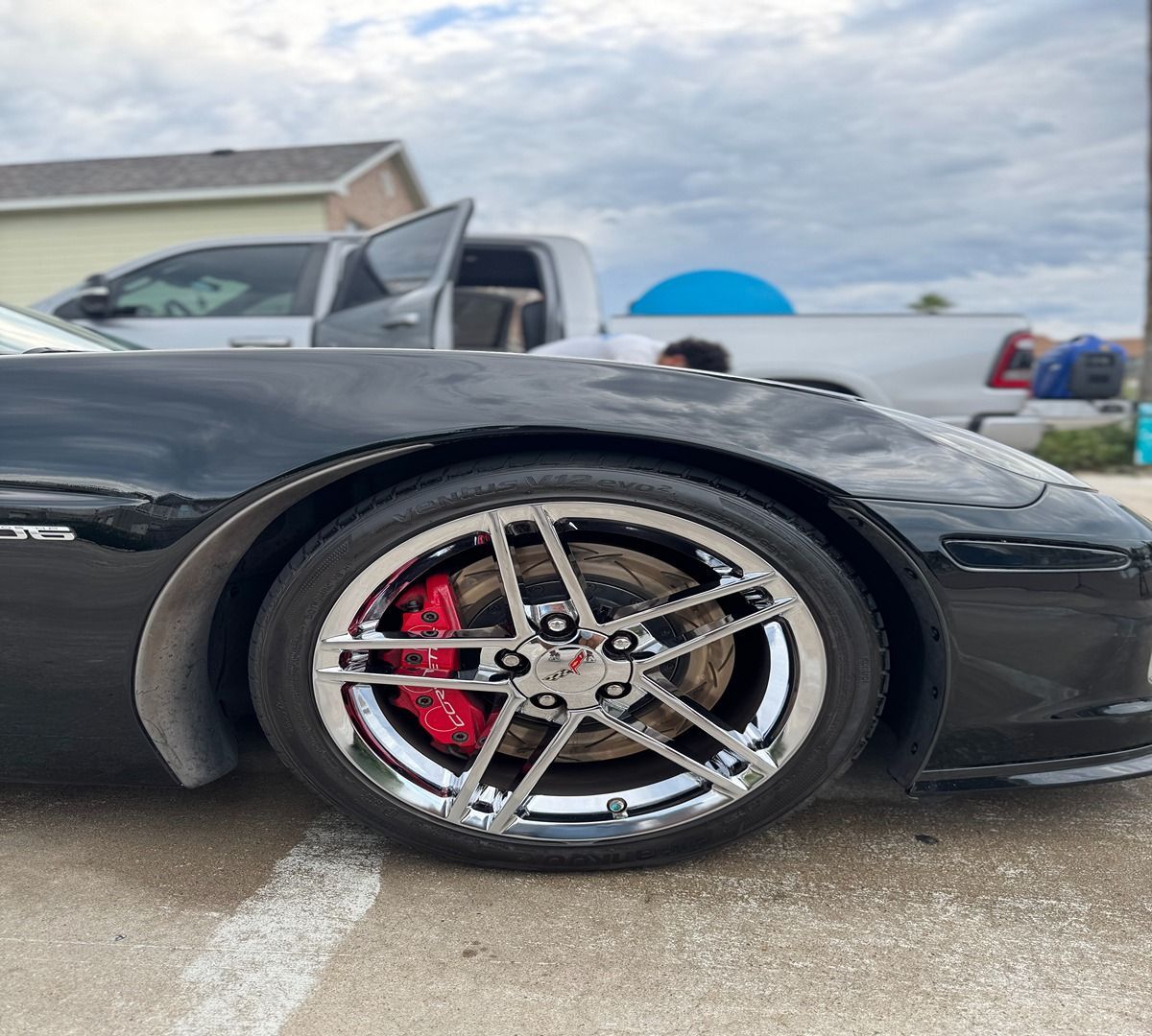 A close-up side view of a black sports car showing its chrome wheel, red brake caliper, and a truck parked in the background.