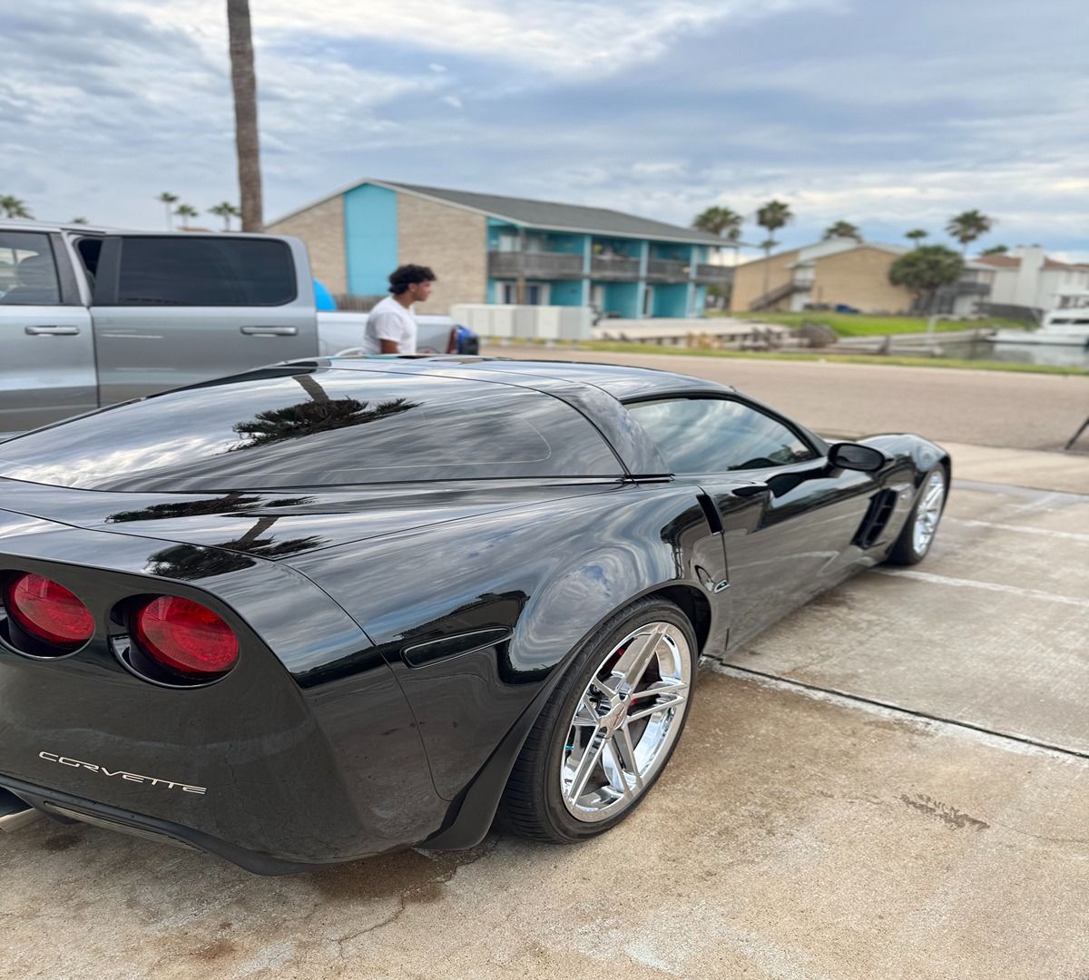A black Chevrolet Corvette parked outdoors next to a silver pickup truck near a multi-story building.