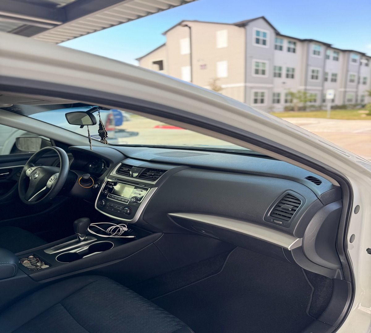Interior view of a parked car showing the dashboard, steering wheel, and front seats with an apartment building outside.