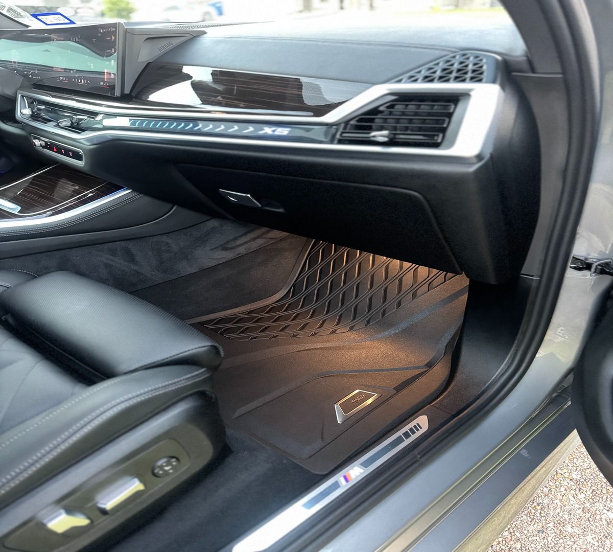The passenger-side interior of a modern BMW featuring black leather seats, a patterned floor mat, and an M-badged trim.