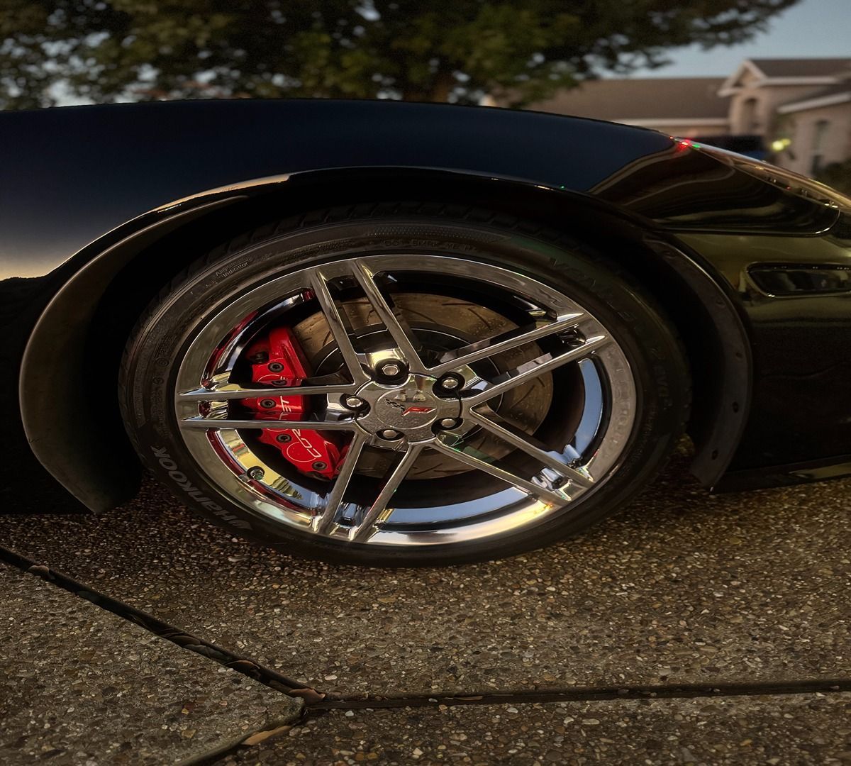 Close-up of a black sports car wheel with chrome spokes and a red brake caliper on a concrete driveway.
