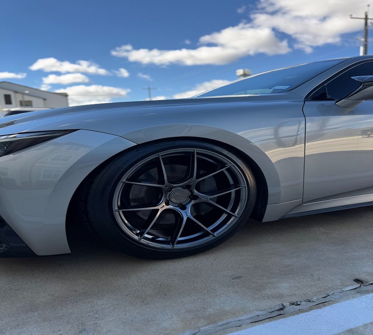 A low-angle view of a silver sedan with custom dark grey multi-spoke wheels parked on concrete under a blue sky.