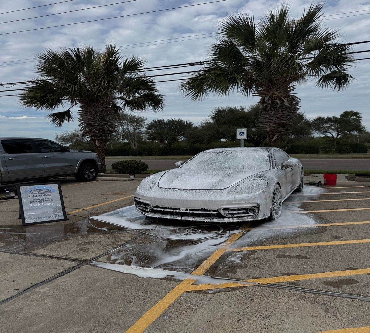 A sports car covered in thick white soap suds parked in a paved lot between two palm trees.