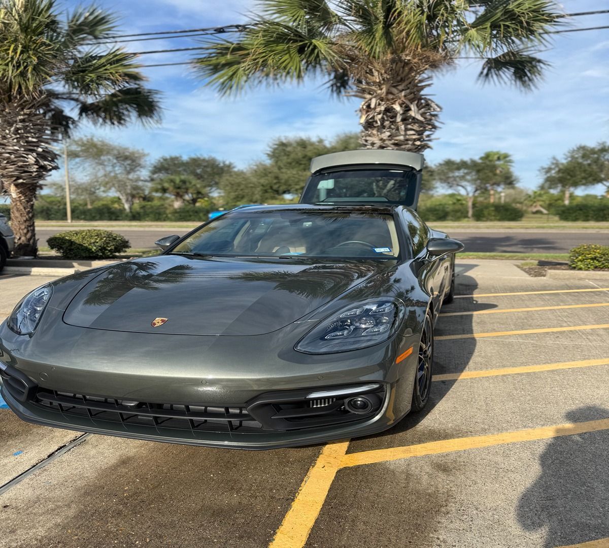 A gray Porsche parked in a sunny lot with its trunk open, framed by palm trees.