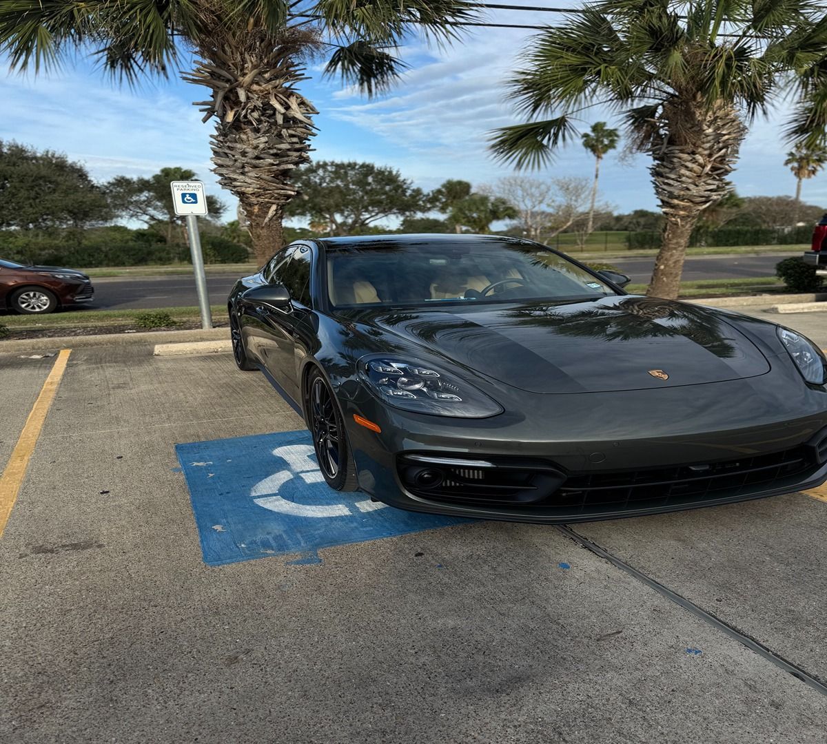 A dark gray Porsche parked in a blue-marked handicapped parking spot outdoors under palm trees.