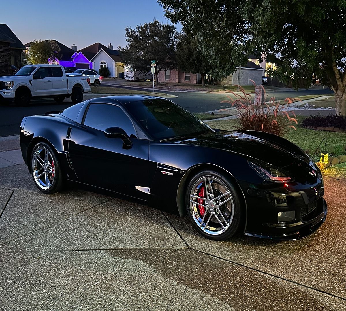 A glossy black Chevrolet Corvette sports car parked on a suburban driveway during sunset, with a white truck in the back.