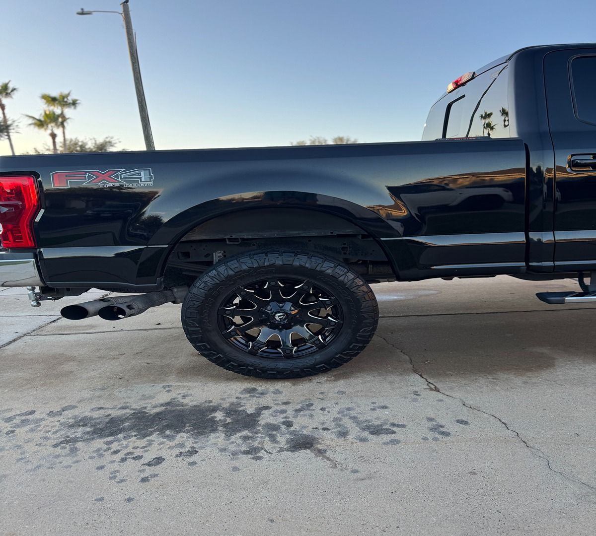 A black Ford truck, showing the rear driver's side fender, tail light, large off-road tire, and dual exhaust pipes.