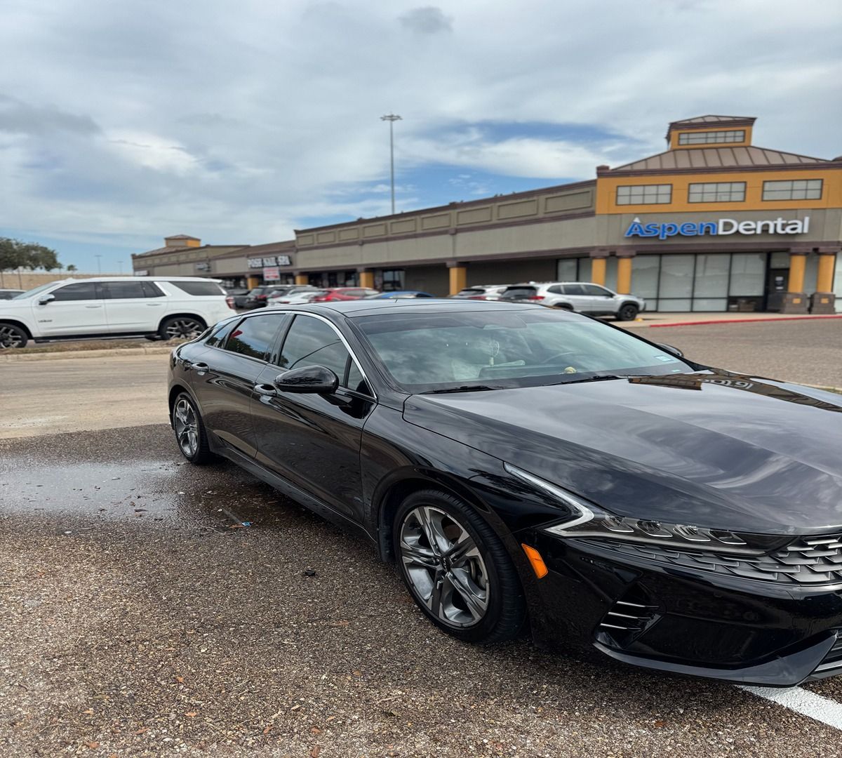 A black Kia K5 parked in front of an Aspen Dental retail center under a cloudy sky.