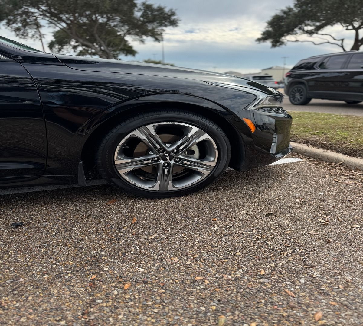 Close-up side view of the front wheel and fender of a parked black car on a gravel lot under a cloudy sky.
