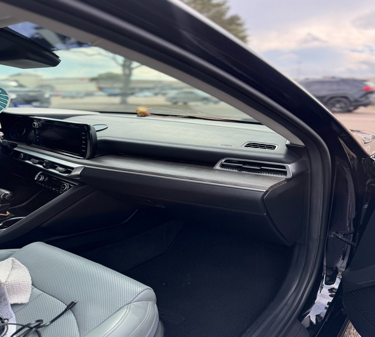 A view from inside a car looking at the dark dashboard, textured gray passenger seat, and an outdoor parking lot.