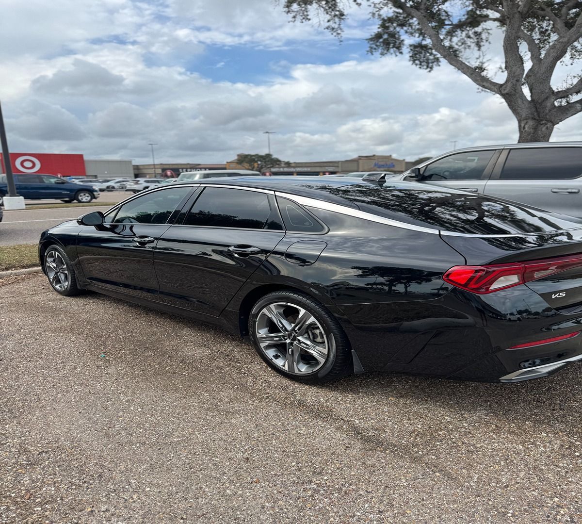 A sleek black Kia K5 sedan parked in a gravel lot with a Target store visible in the background.