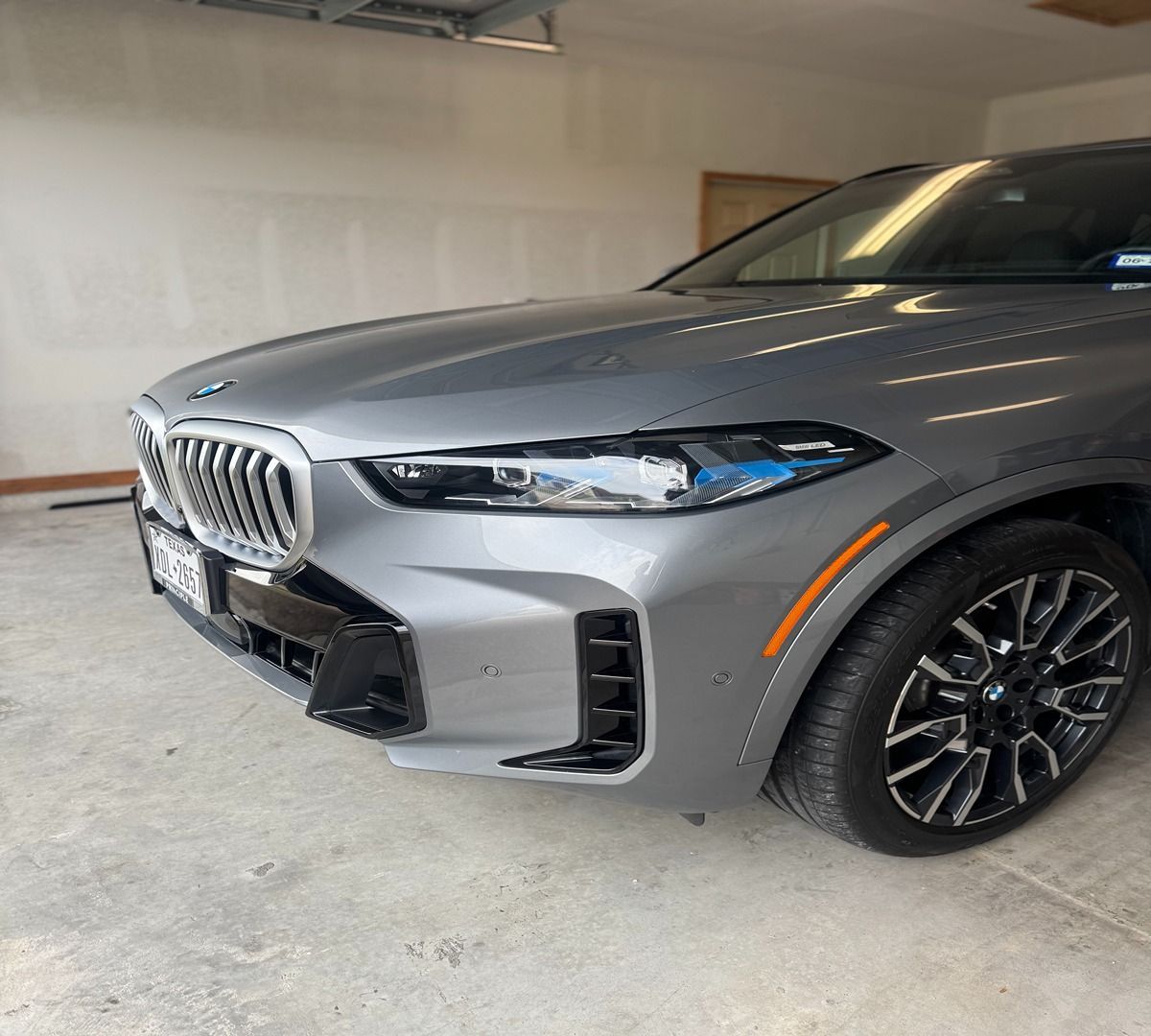 Front view of a gray BMW SUV parked in a garage, focusing on the headlight, grille, and front wheel.