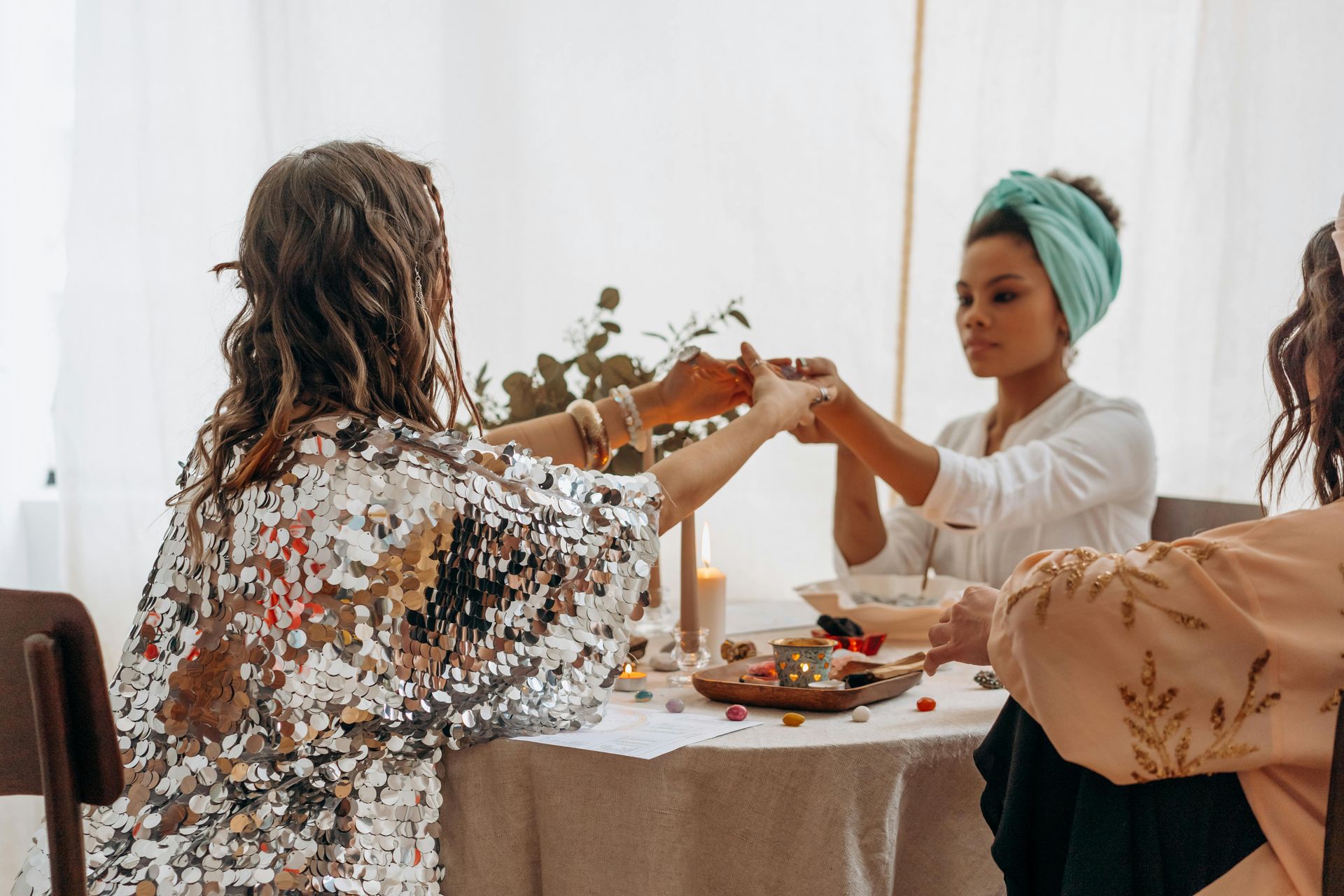 Three people holding hands at a table, likely in prayer. Silver sequined top, turquoise head wrap, candles.