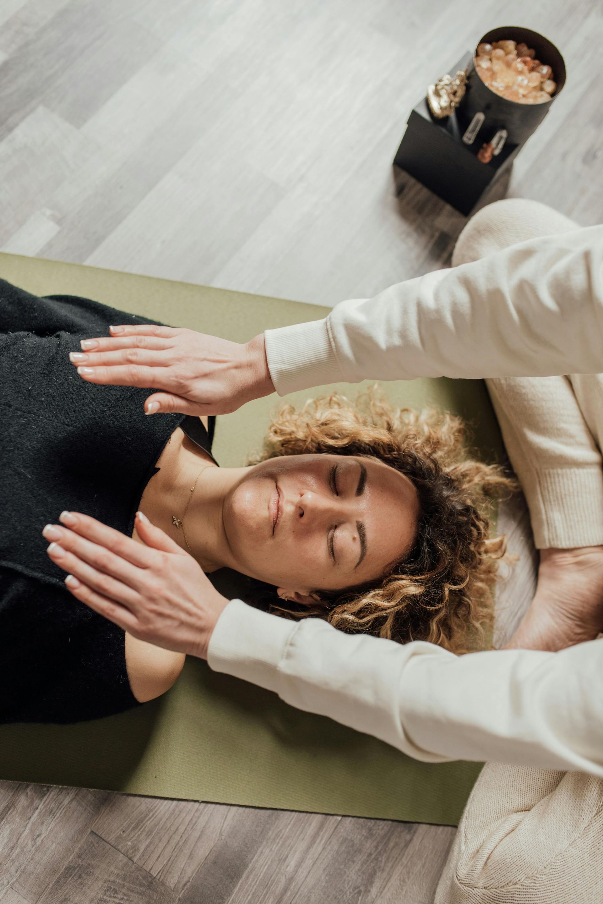 Hands of two people over the chest of a woman lying on a mat, eyes closed, receiving energy healing.