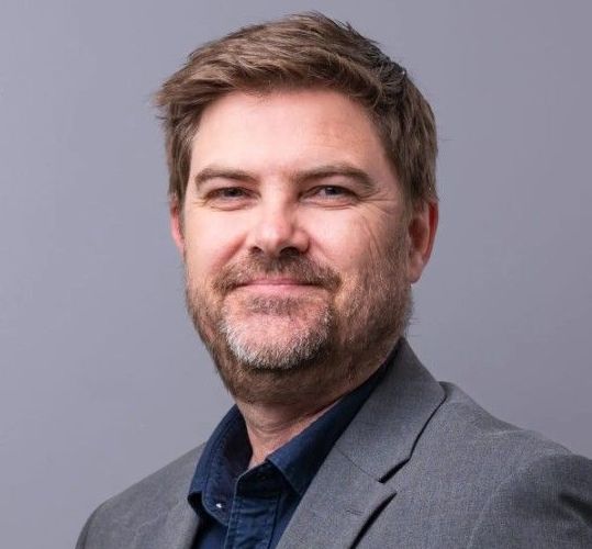 Man with a beard, wearing a grey blazer over a dark blue shirt, smiling at the camera.