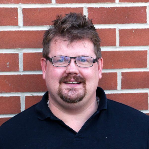 Man with glasses and goatee smiling in front of a brick wall. He wears a black polo shirt.