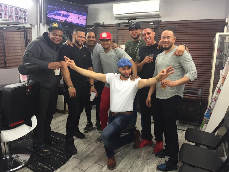 Group of men smiling in a barbershop, posing together around a seated man in a blue bandana.