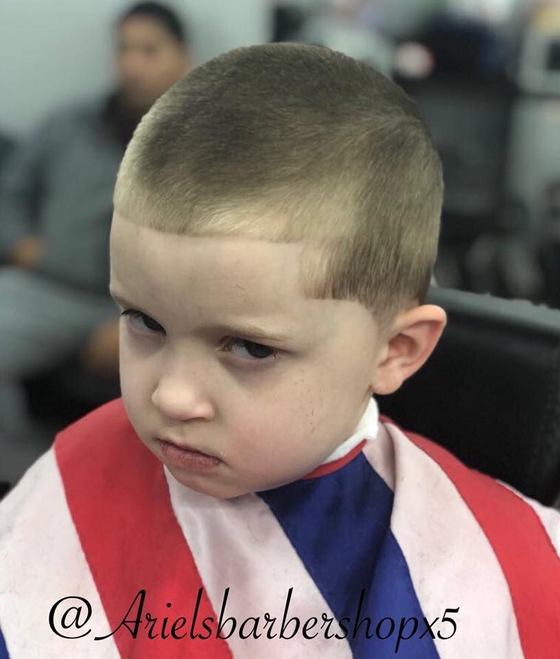 Child with a short buzz cut and serious expression wearing a red, white, and blue barber cape.