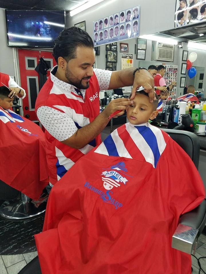 Barber trimming a client’s hair in a red cape at a shop, with mirrors and wall posters behind them.