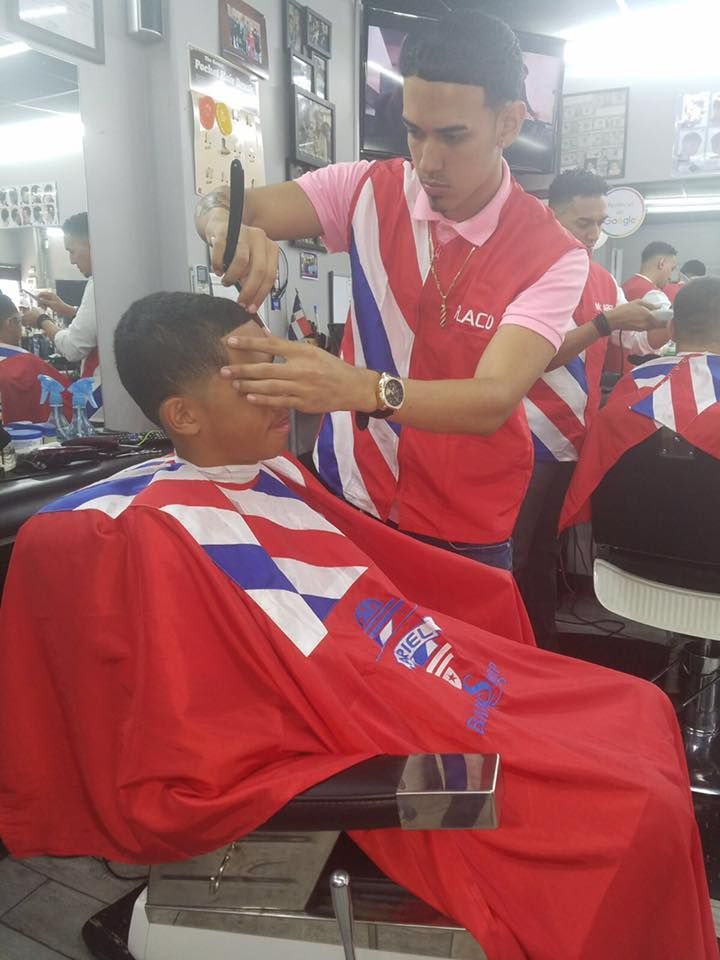 Barber in red shirt trims a client’s hair in a busy barbershop.