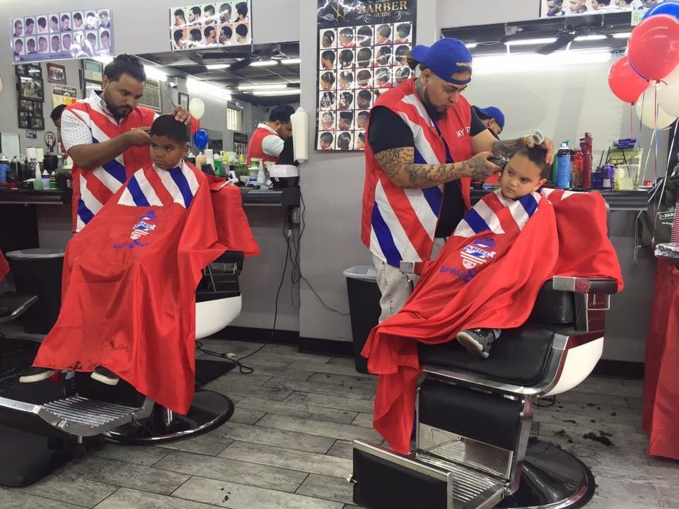 Barbers giving haircuts to two seated clients in red capes inside a busy barbershop