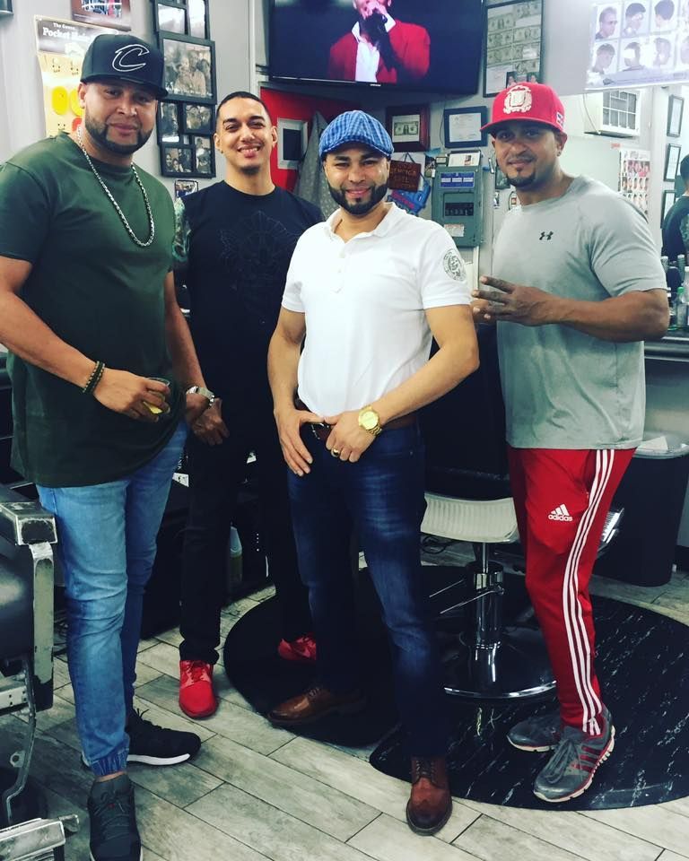 Four men posing together in a barbershop, with TVs and chairs in the background.
