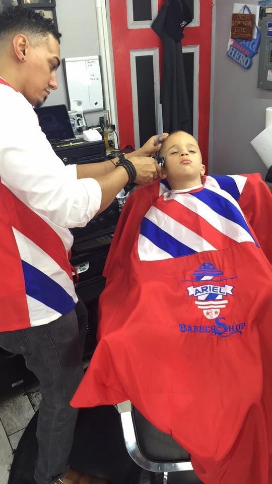 Barber trims a client’s hair in a shop, both wearing red-white-blue capes.