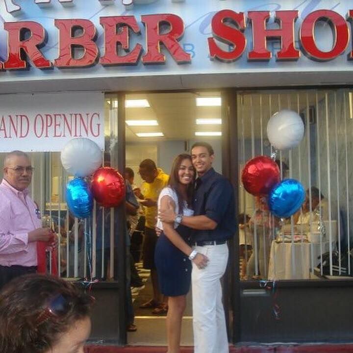 Couple posing outside a barber shop at a grand opening, with red and blue balloons.